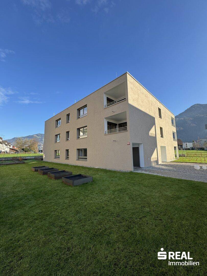 Modernes Mehrfamilienhaus mit Balkonen, umgeben von einer gepflegten Grünfläche und Bergblick.