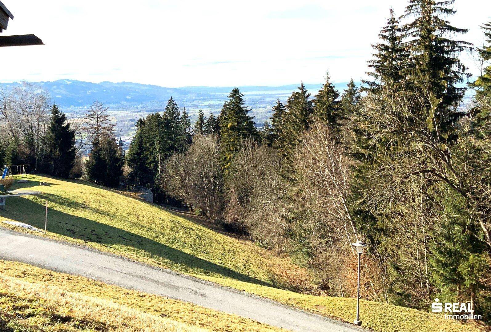 Panoramablick über grüne Hügel, Wälder und einen See in der Ferne, mit einem kleinen Spielplatz.