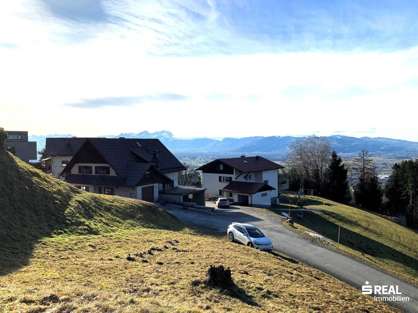 Panoramablick auf Wohnhäuser in hügeliger Landschaft mit Bergen im Hintergrund unter blauem Himmel.