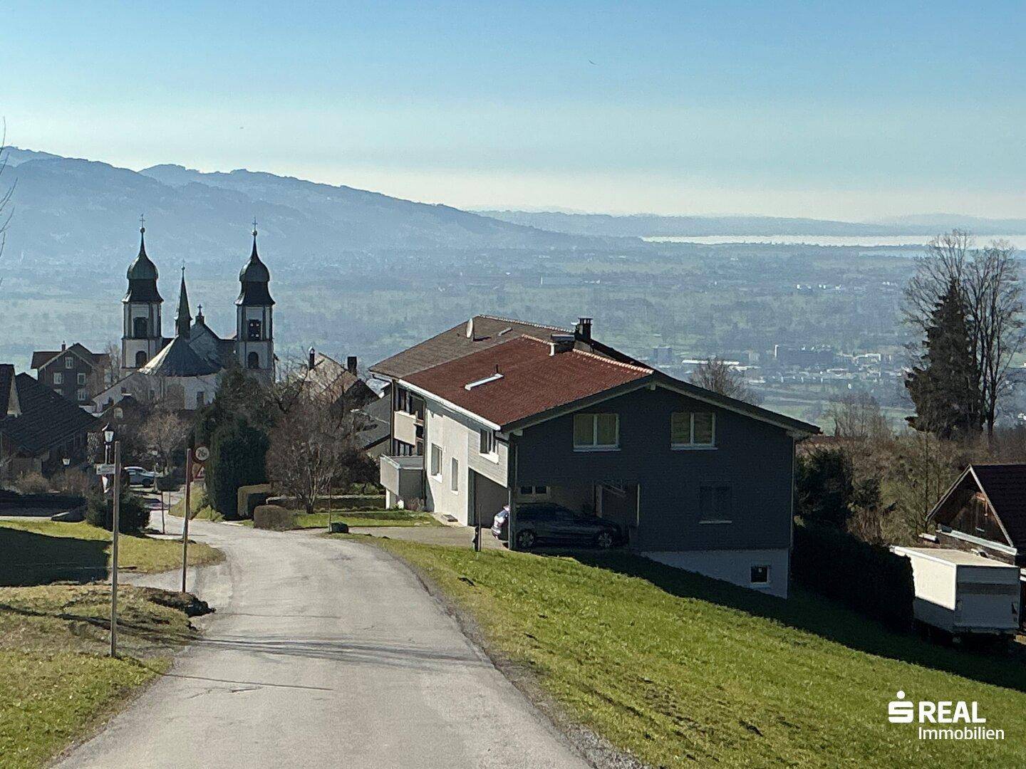 Wohnhaus in Hanglage mit weitem Blick über das Tal, Berge und See im Hintergrund.