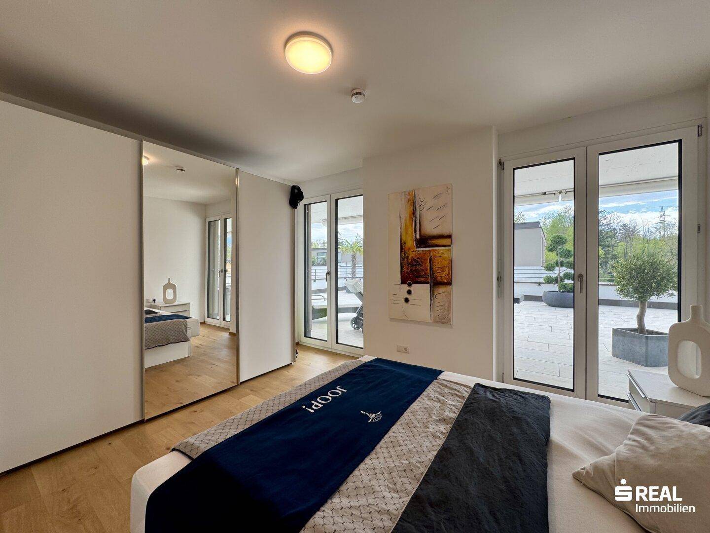 Schlafzimmer mit Spiegelschränken, Holzboden und großen Fenstern zur Terrasse.