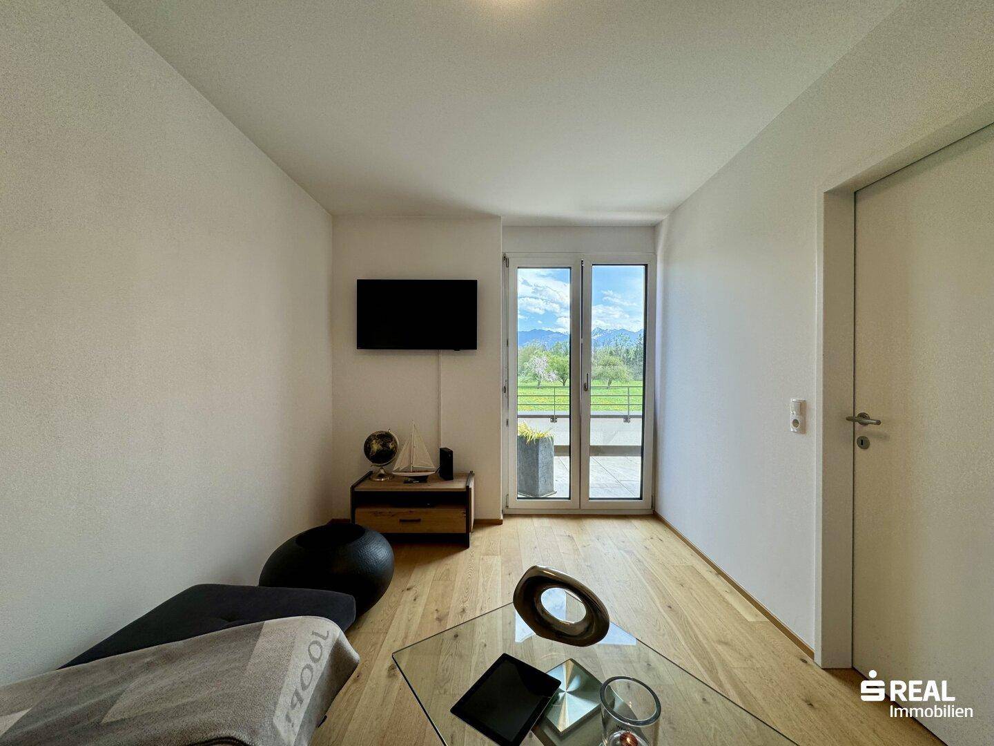 Helles Wohnzimmer mit Fernseher, Holzboden und Zugang zu einem kleinen Balkon.