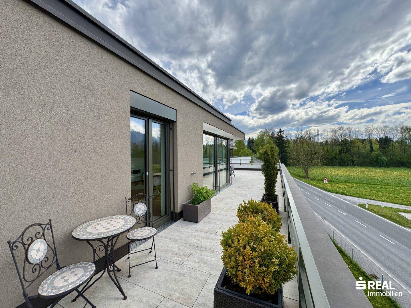 Seitlicher Terrassenbereich mit kleinem Tisch, Pflanzen und Blick auf die Straße und Landschaft.