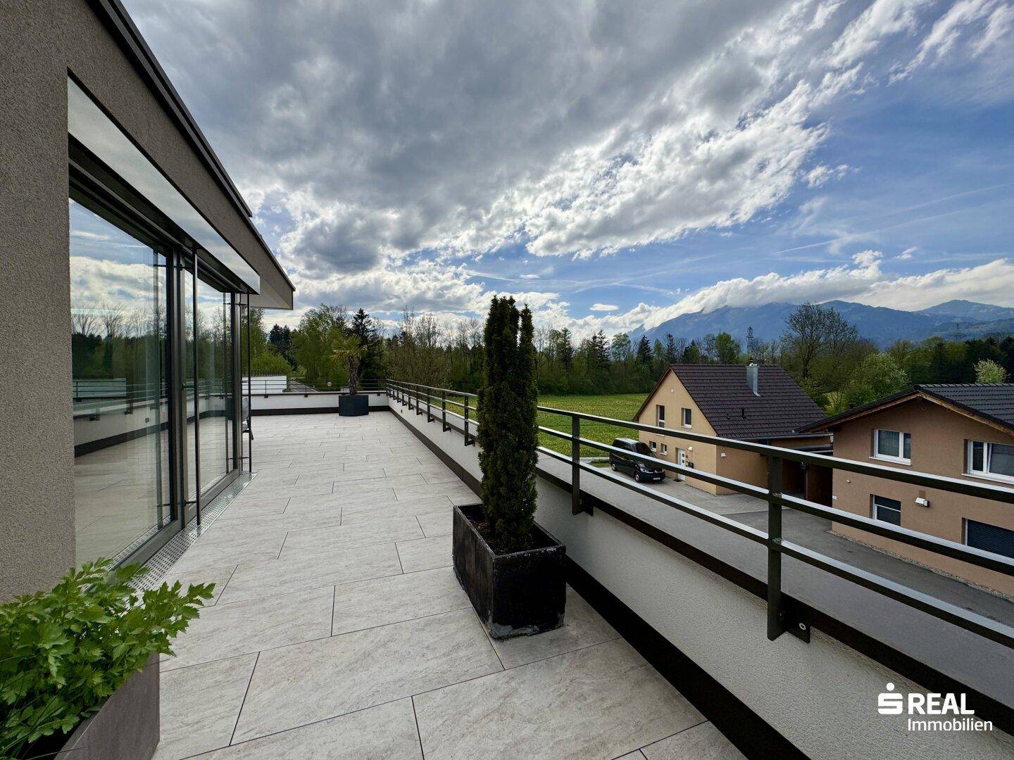 Lange Terrasse mit Metallgeländer, Pflanzen und weitem Blick auf die grüne Umgebung.