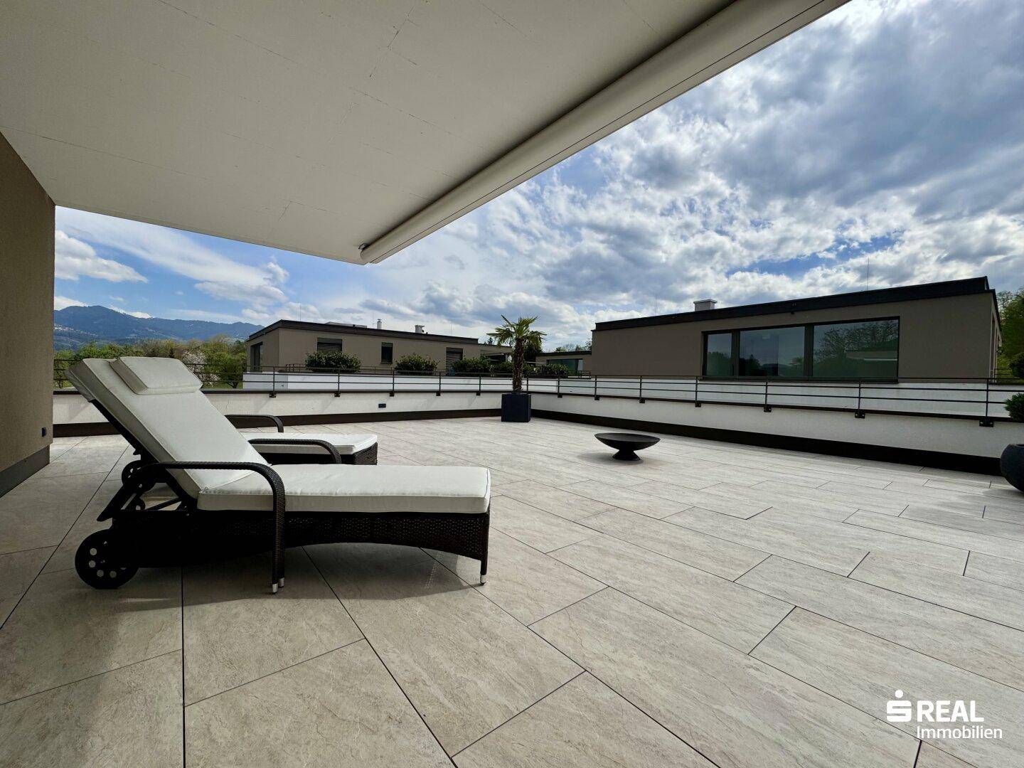 Sonnige Dachterrasse mit Liegestühlen und Blick auf die benachbarten Gebäude und Berge.