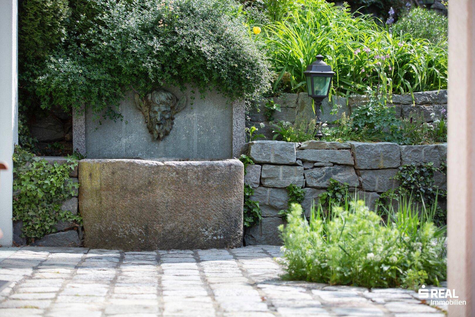 Historischer Steinbrunnen mit kunstvoller Verzierung und Natursteinmauer im grünen Gartenbereich.