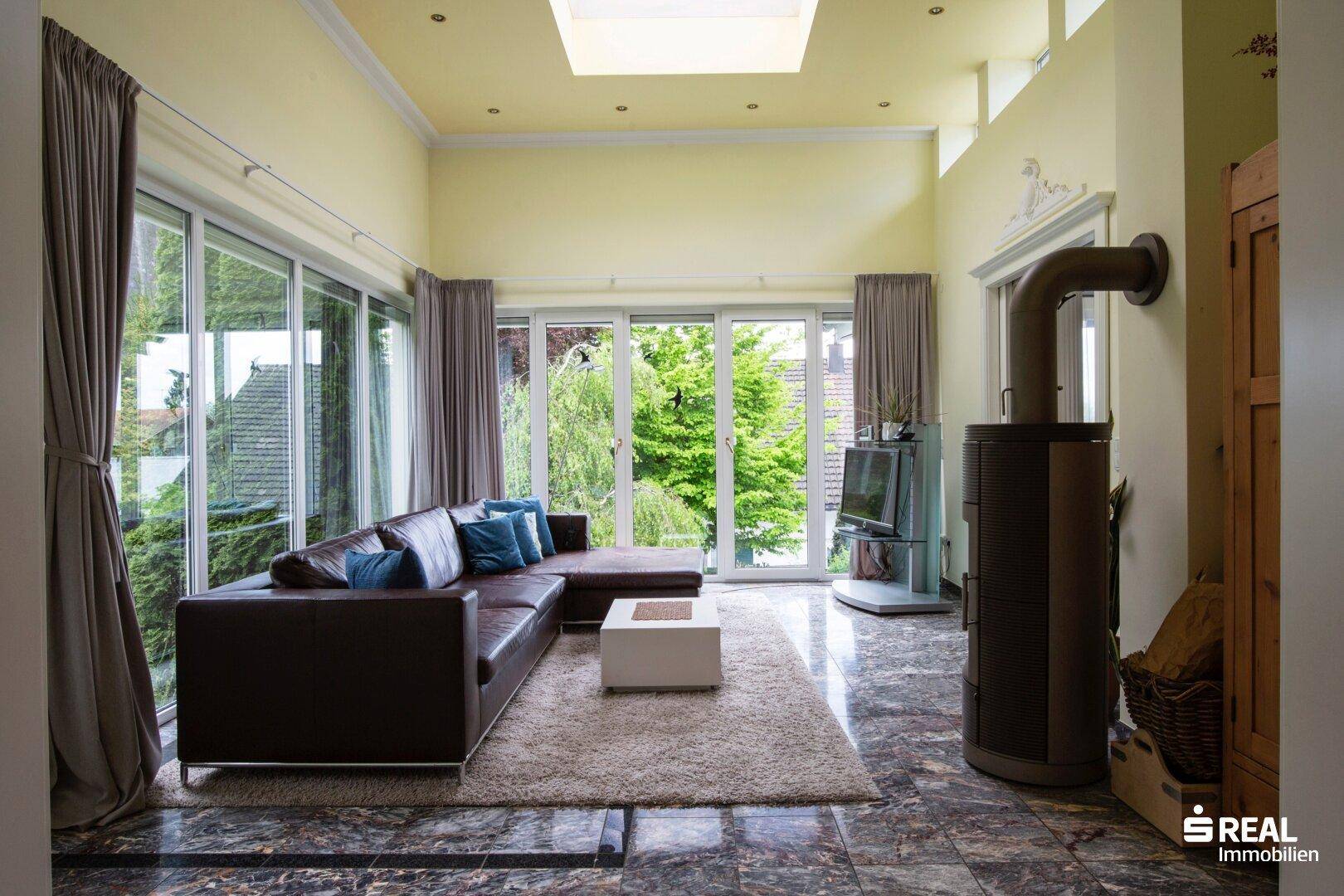 Lichtdurchflutetes Wohnzimmer mit Ecksofa, großen Fenstern, Dachfenster und modernem Kaminofen.