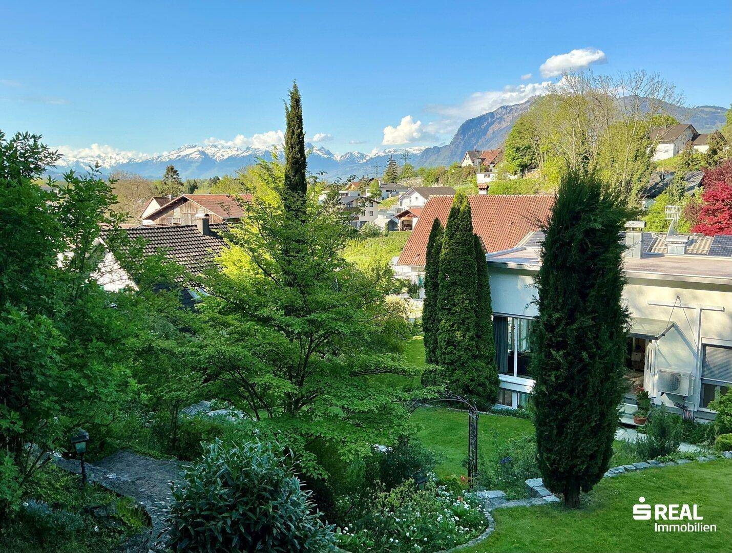 Panoramablick auf die umliegende Wohngegend mit grünen Gärten und majestätischen Bergen im Hintergrund.