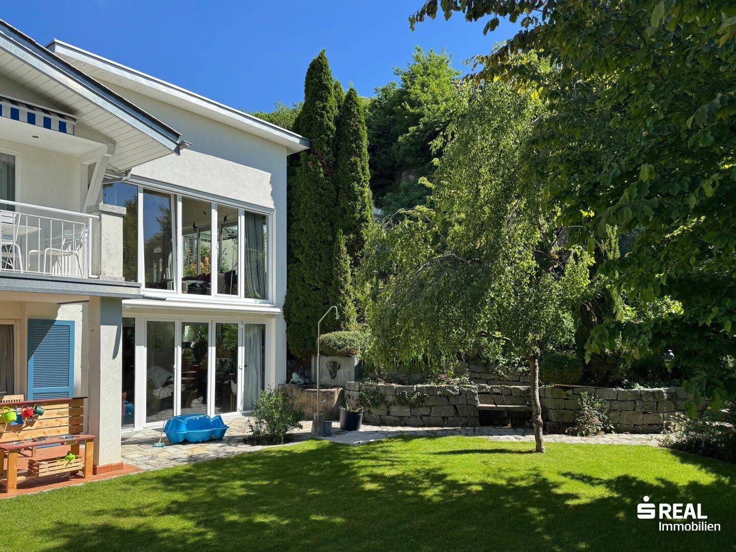 Heller Gartenbereich mit Rasenfläche, hohen Bäumen und moderner Hausfassade mit großen Fenstern.