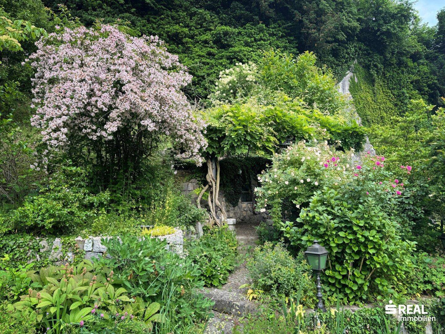 Üppiger, blühender Garten mit verschlungenen Wegen und einer von Pflanzen bewachsenen Pergola.