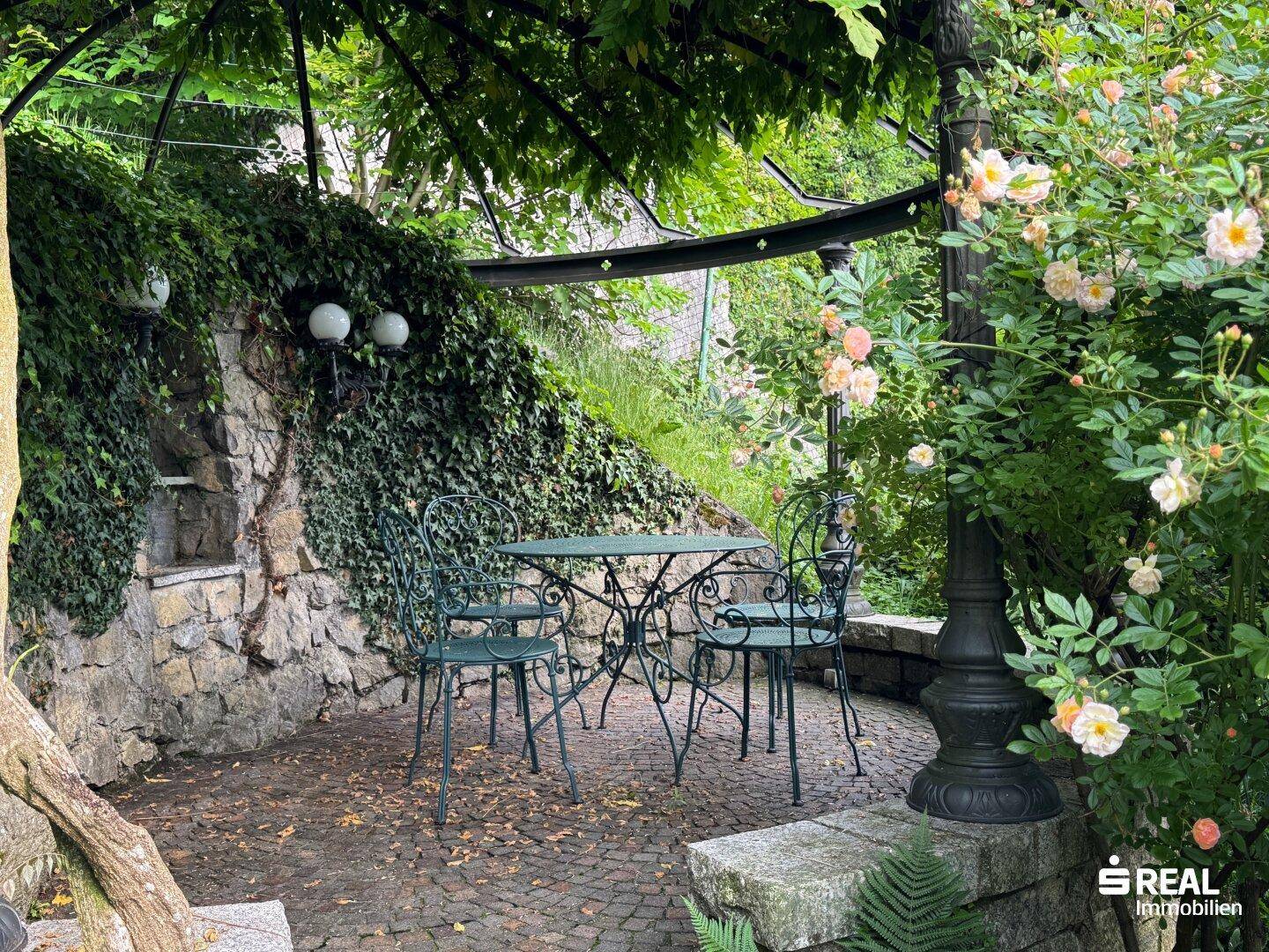 Romantischer Sitzbereich im Garten unter einer Pergola, umgeben von Rosen und Steinmauern.