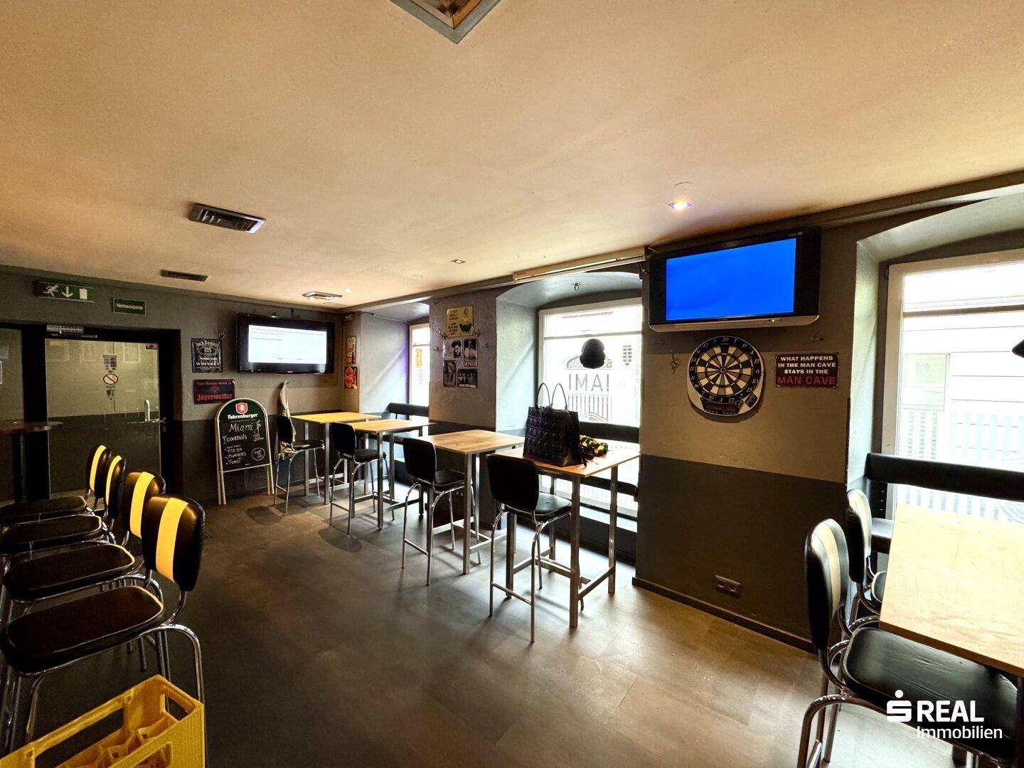 Sitzbereich einer Bar mit Tischen, Stühlen und einem Fernseher, gut beleuchtet durch Fenster.