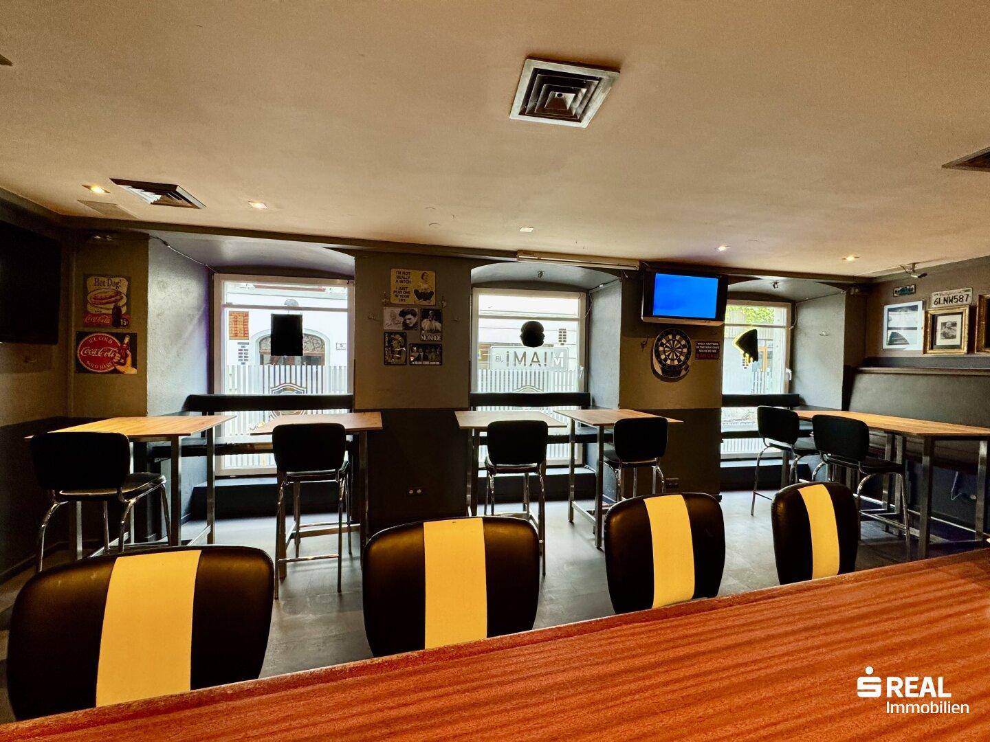 Innenansicht einer Bar mit Sitzgelegenheiten, Tischen und einem Fernseher an der Wand, mit Tageslicht.