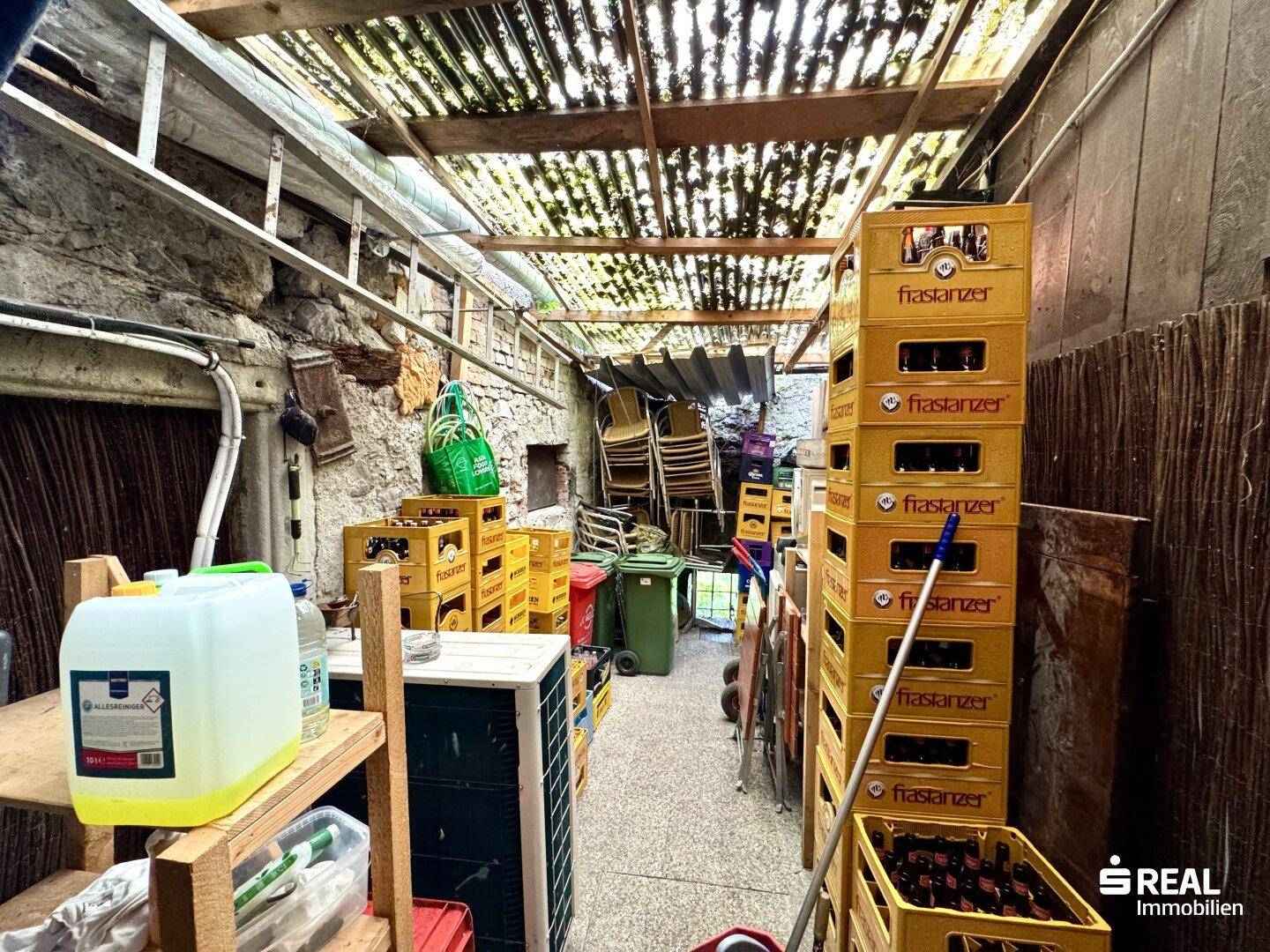 Überdachter Lagerbereich im Freien mit gestapelten Getränkekisten und verschiedenen Gegenständen.