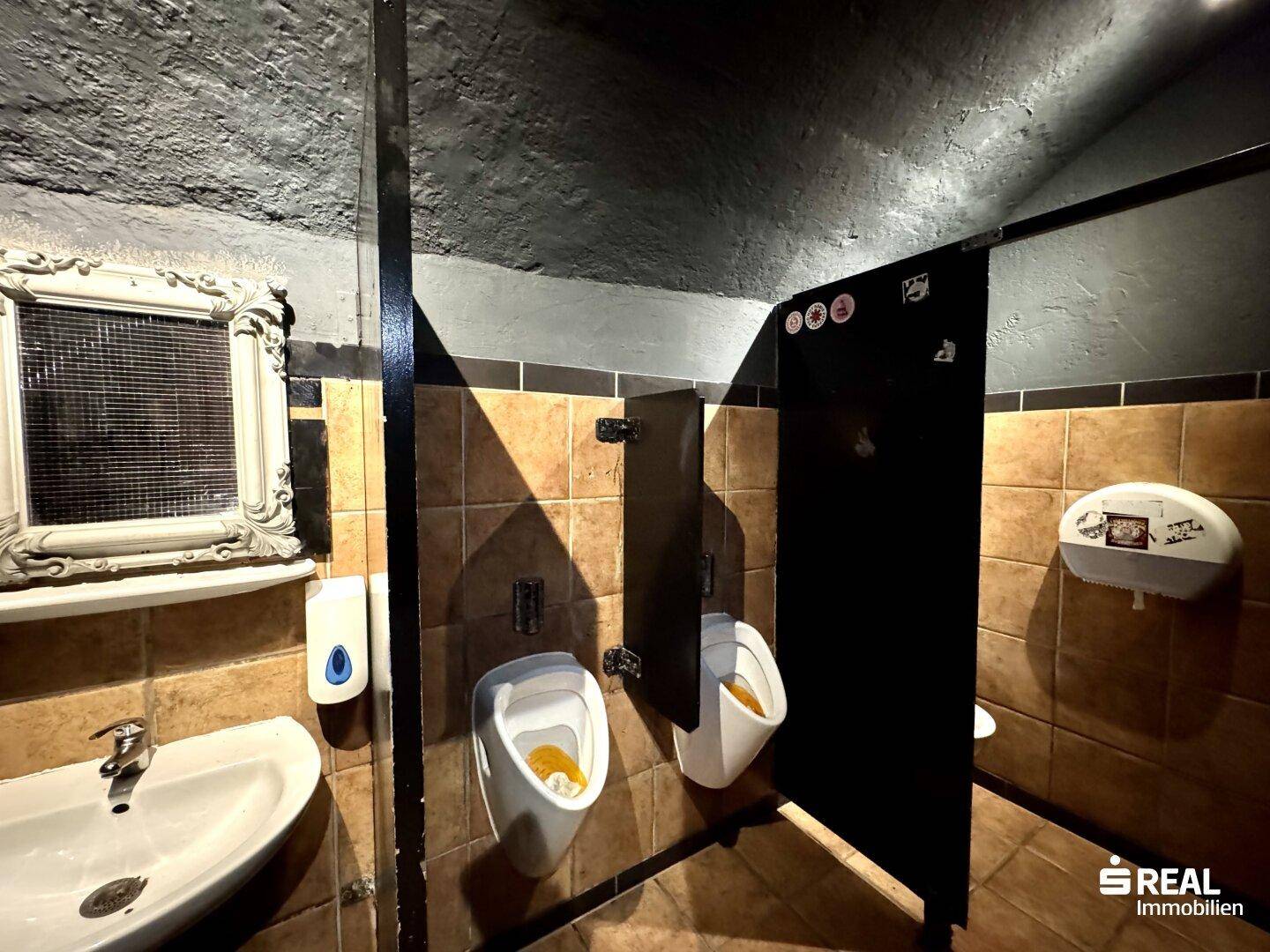 Herren-WC mit Urinalen, Waschbecken und gefliesten Wänden in einem rustikalen Stil.