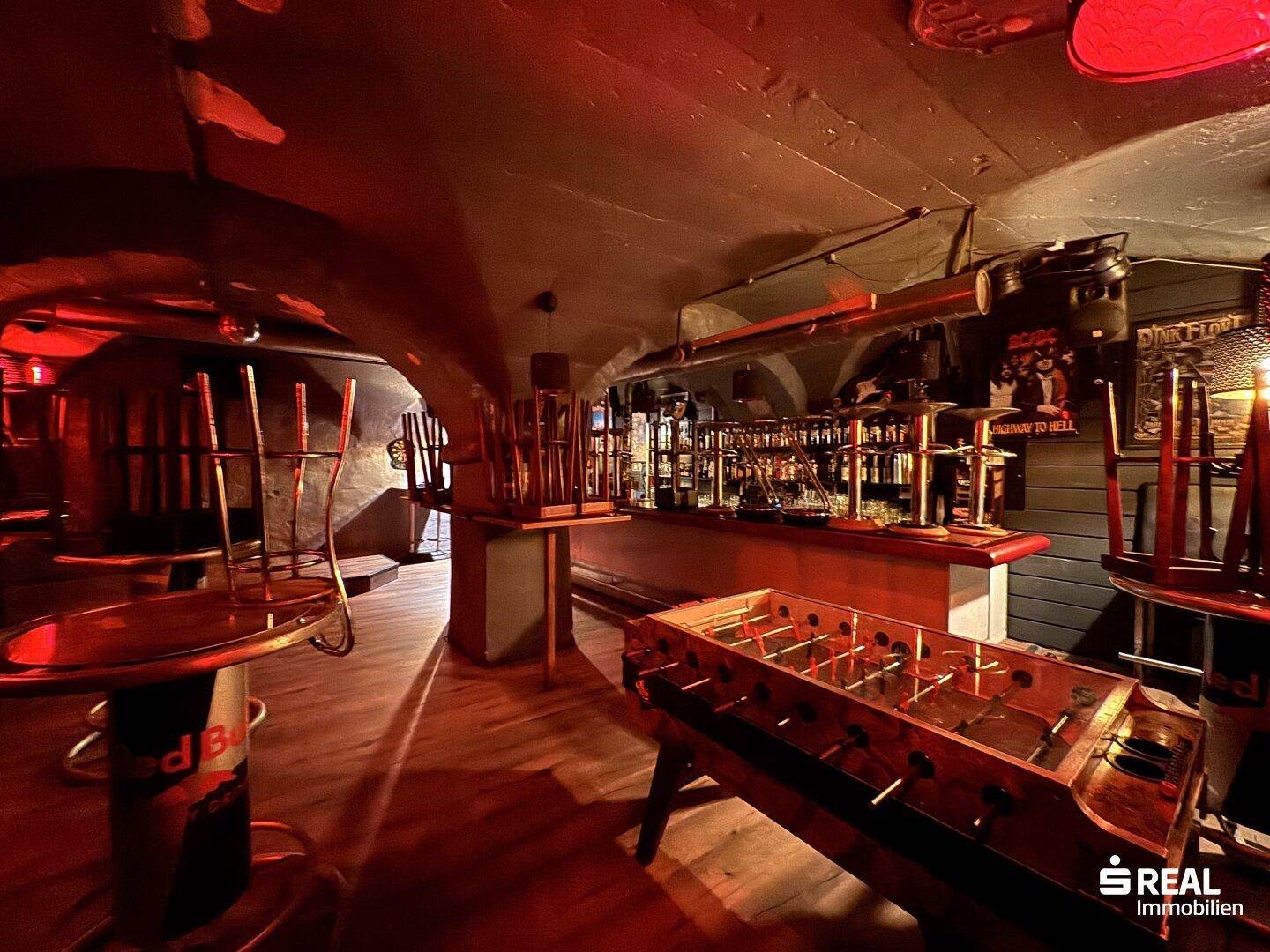 Kellerbar mit roter Beleuchtung, Holzboden und einem Tischfußballspiel, rustikale Atmosphäre.