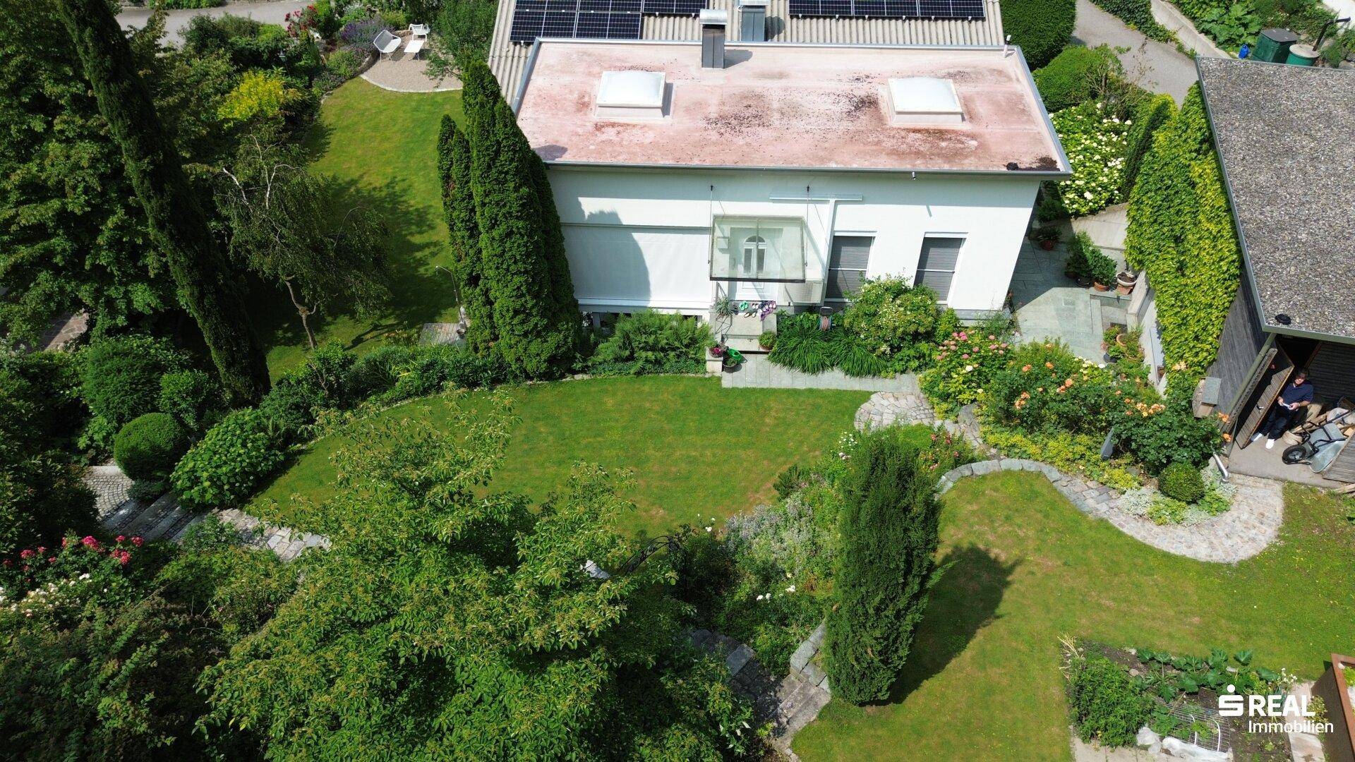 Rückansicht des Hauses mit sonnigem Garten, Photovoltaikanlage und einem kleinen Gewächshaus.