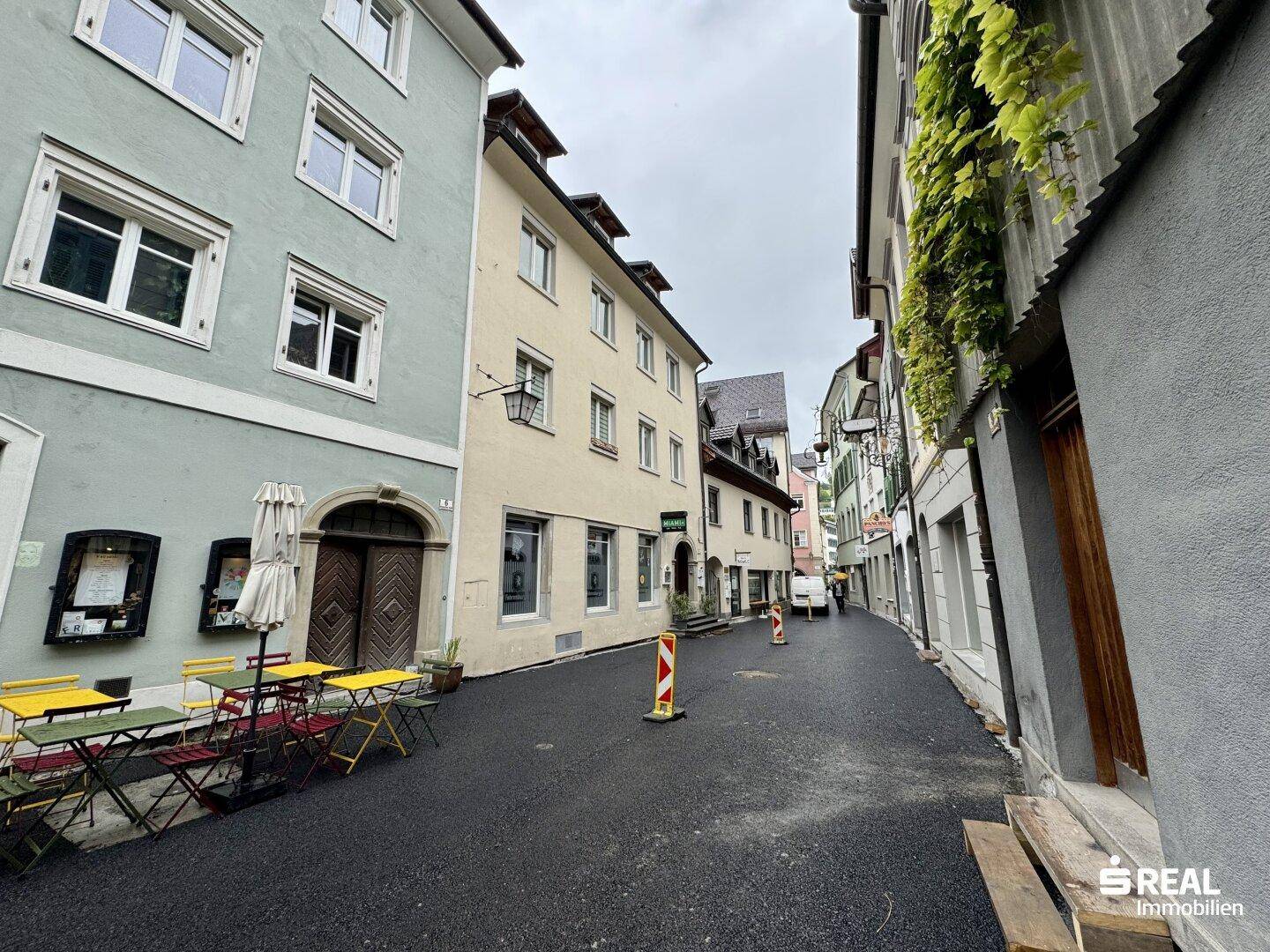 Historische Gasse mit Gebäuden, Außengastronomie und Kopfsteinpflaster, belebte Atmosphäre.