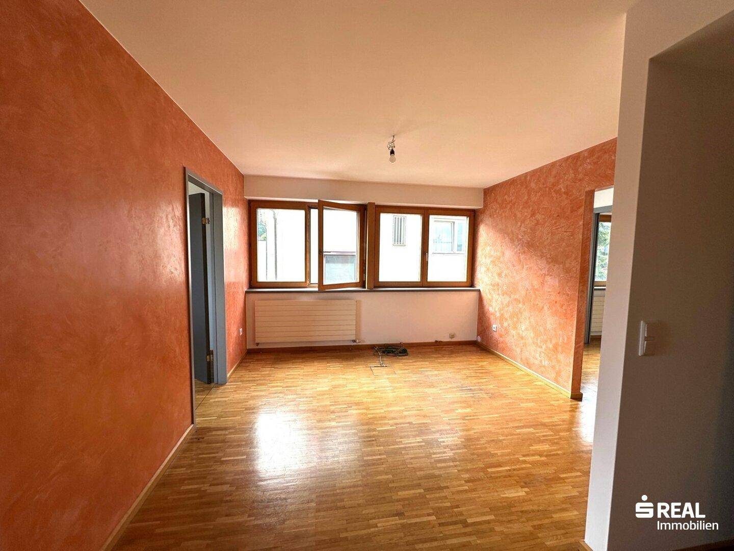 Heller Wohnraum mit Holzboden, großen Fenstern und einer markanten orangefarbenen Wandgestaltung.