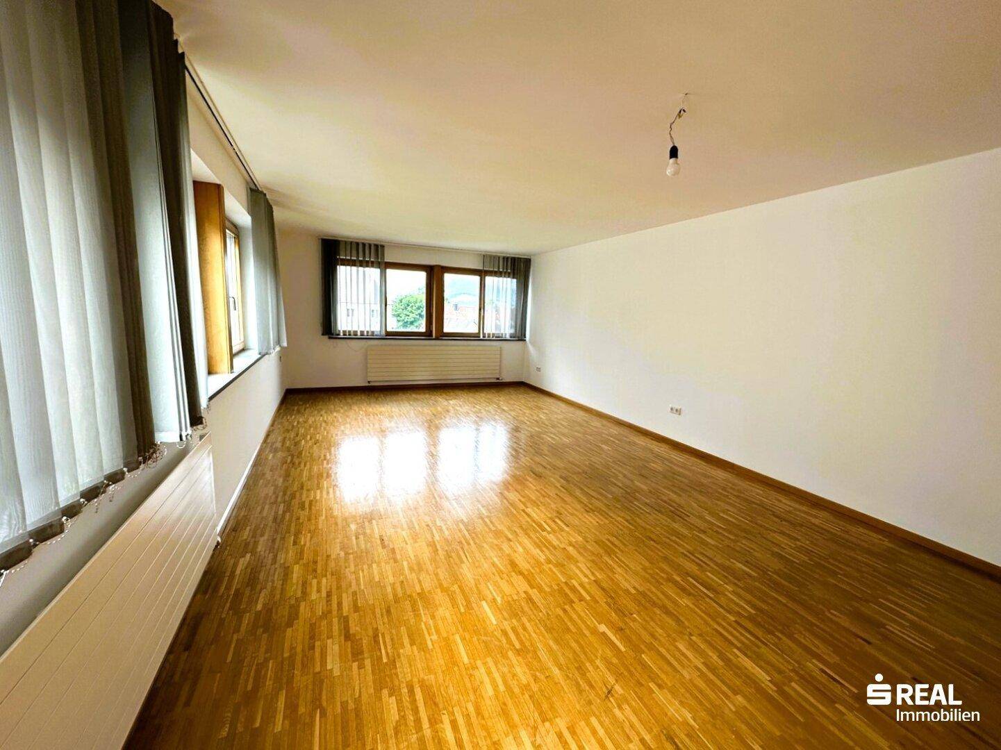 Geräumiger Wohnbereich mit viel Tageslicht, Holzparkett und Jalousien an den großen Fenstern.