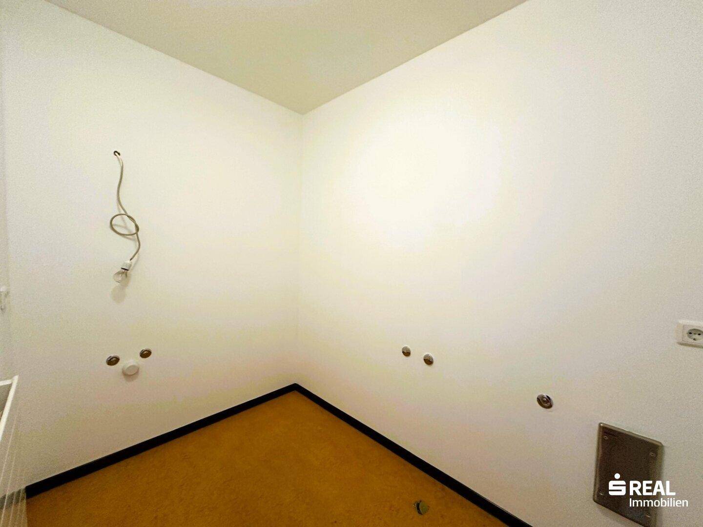 Leerer Raum mit gelbem Teppichboden und Anschlüssen an der Wand, möglicherweise für eine Waschküche.
