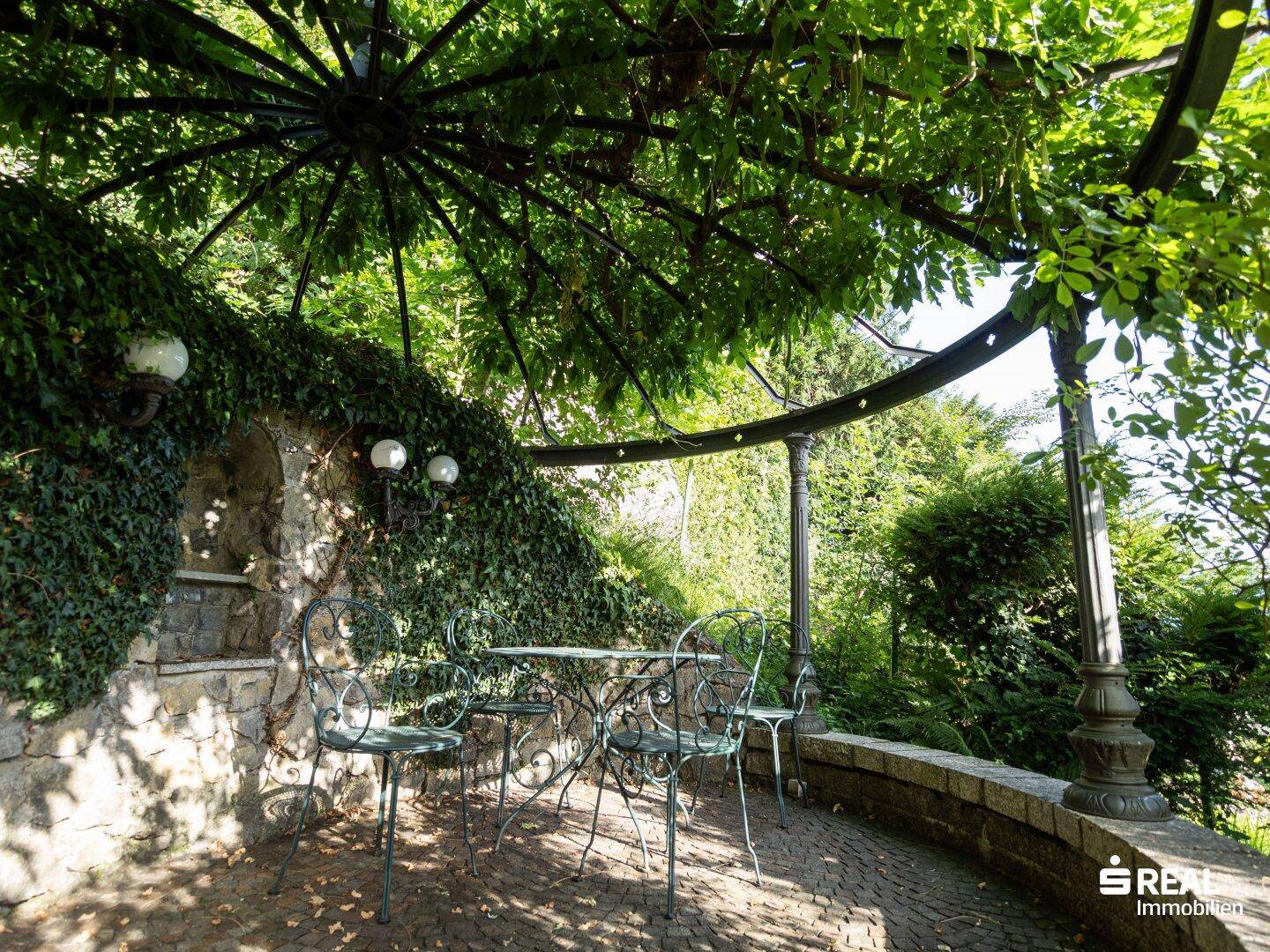 Idyllische Terrasse mit schmiedeeisernen Möbeln unter einer grünen Pergola, umgeben von üppiger Vegetation.