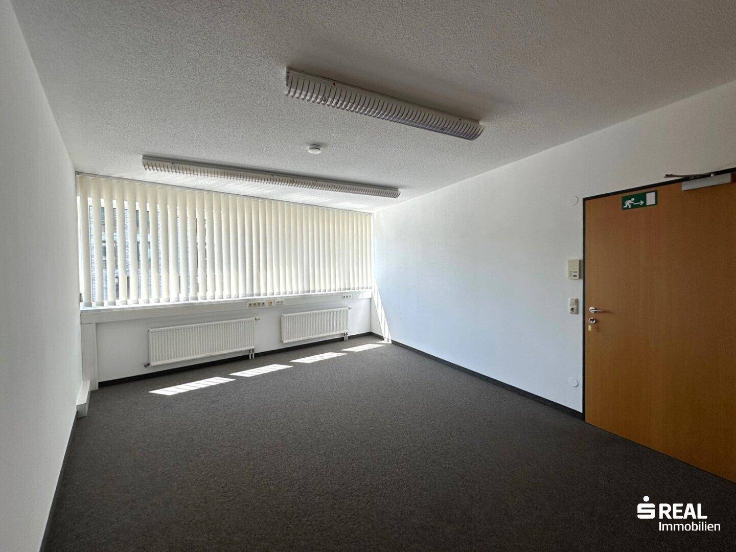 Helles Büro mit Teppichboden, großen Fenstern, Heizkörpern und vertikalen Jalousien für Tageslichtregulierung.