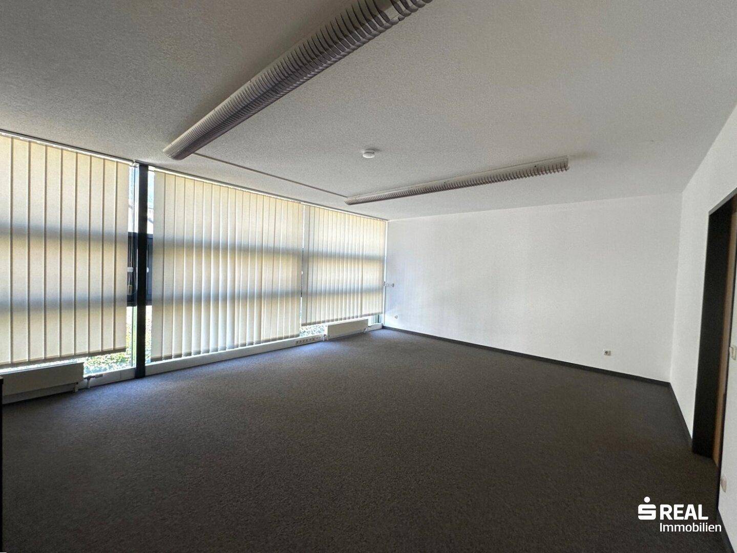 Weitläufiges Büro mit Teppichboden und einer Fensterfront, die viel Tageslicht hereinlässt und Jalousien bietet.