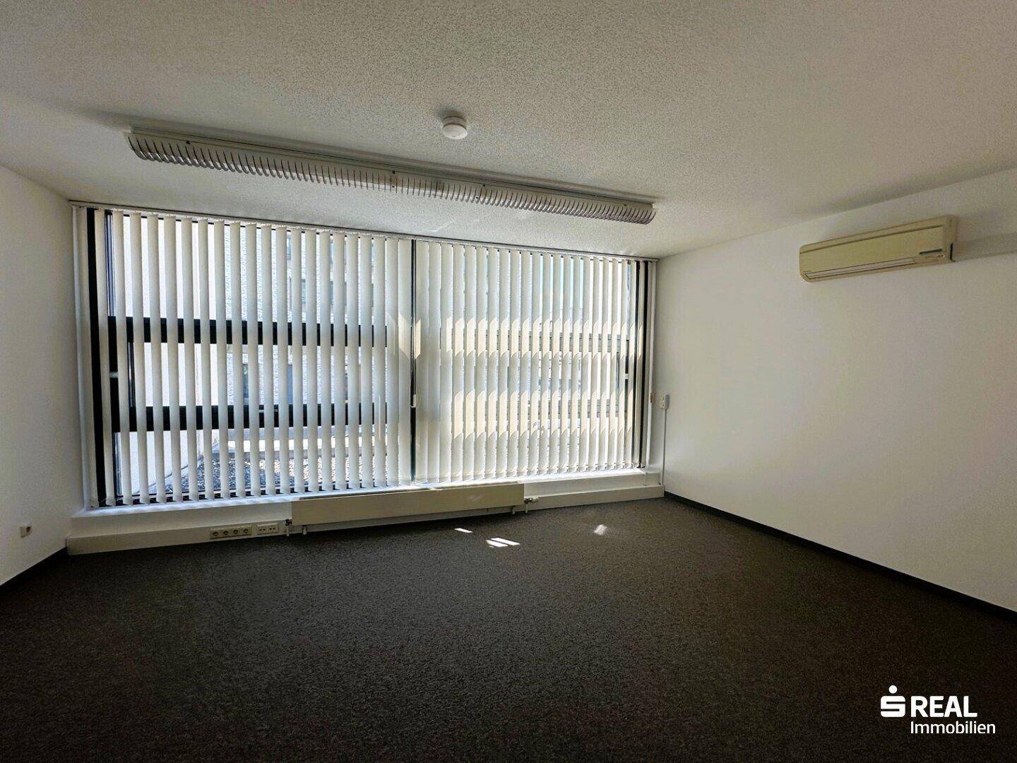 Bürofläche mit Teppichboden, großer Fensterfront mit Jalousien und Klimaanlage an der Wand montiert.