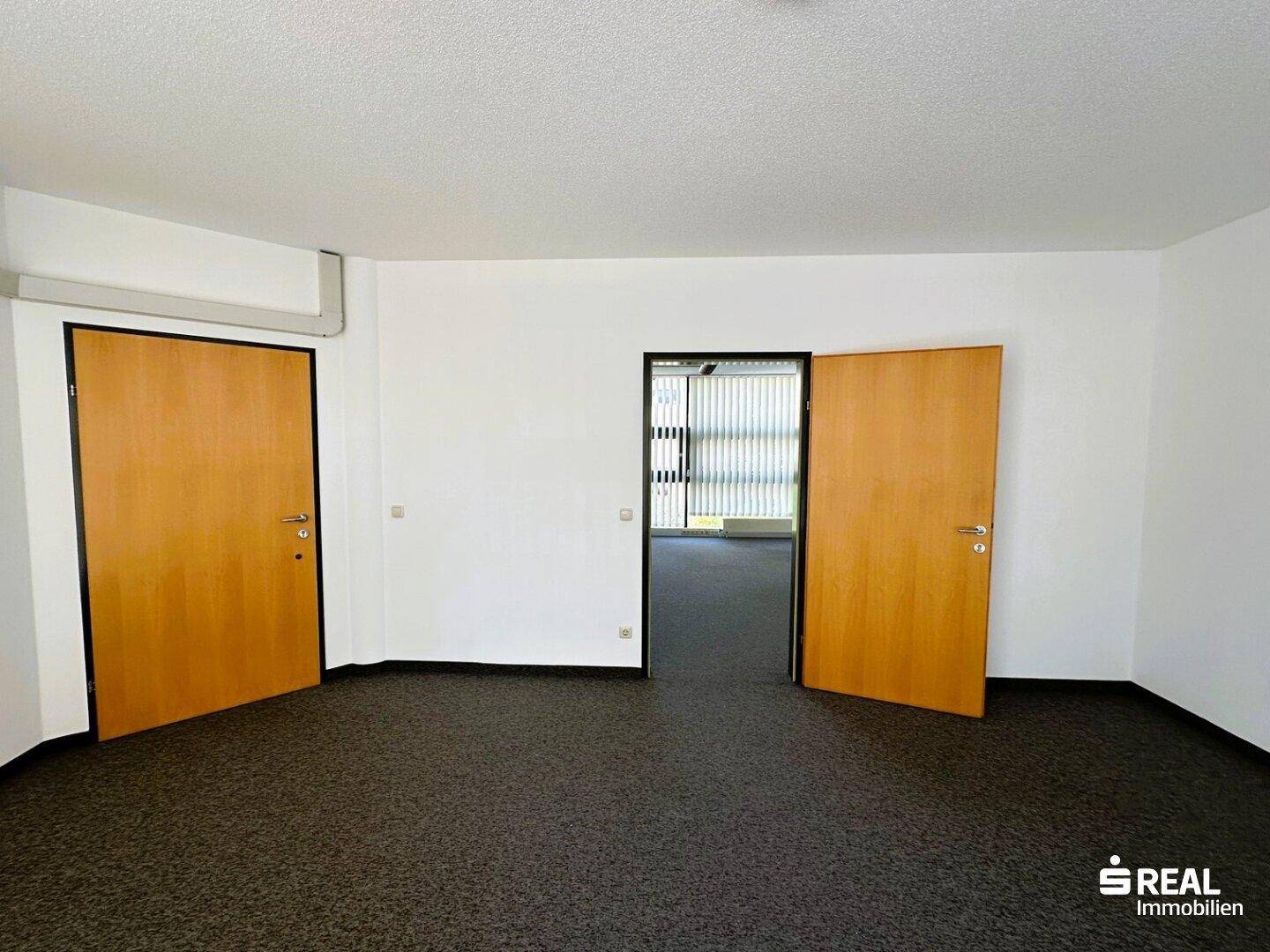 Büro mit Teppichboden und zwei Holztüren, eine davon führt in einen weiteren Arbeitsbereich.