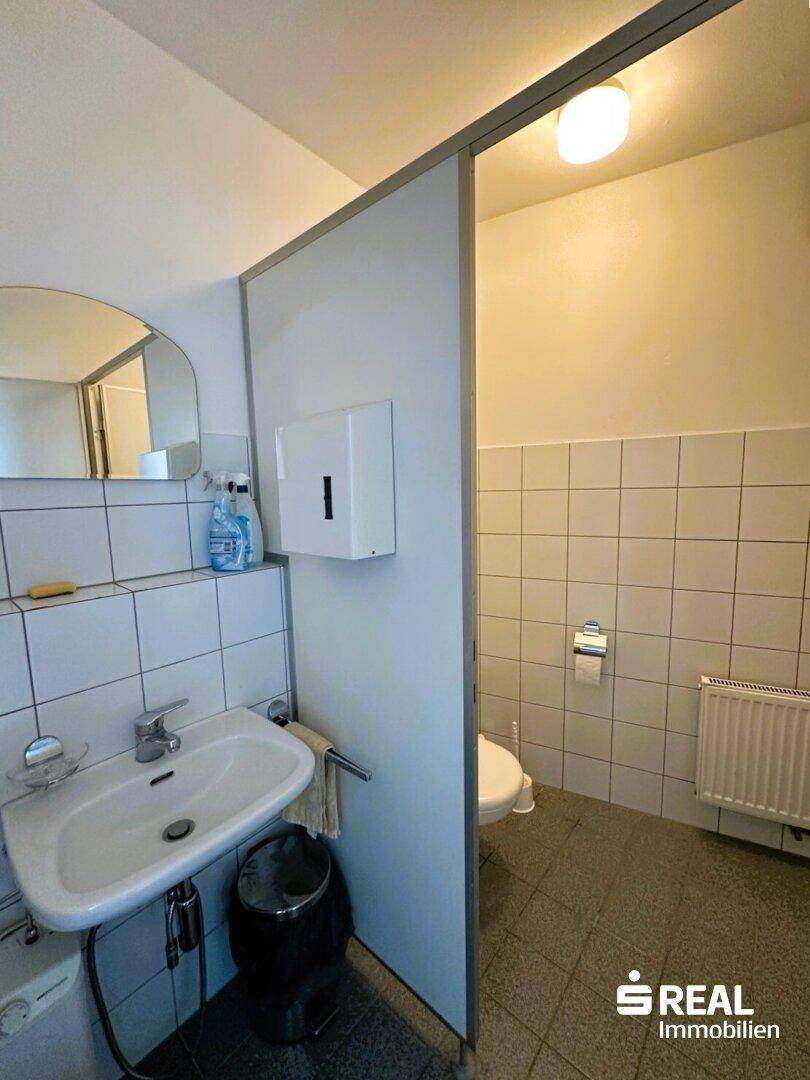 Toilettenraum mit weißen Wandfliesen, Waschbecken, Spiegel und Toilette hinter einer Trennwand.