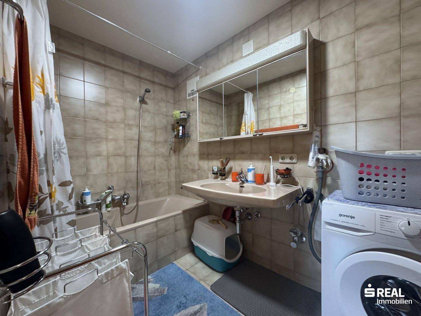 Gefliestes Badezimmer mit Badewanne, Waschbecken, Spiegelschrank und Waschmaschine.