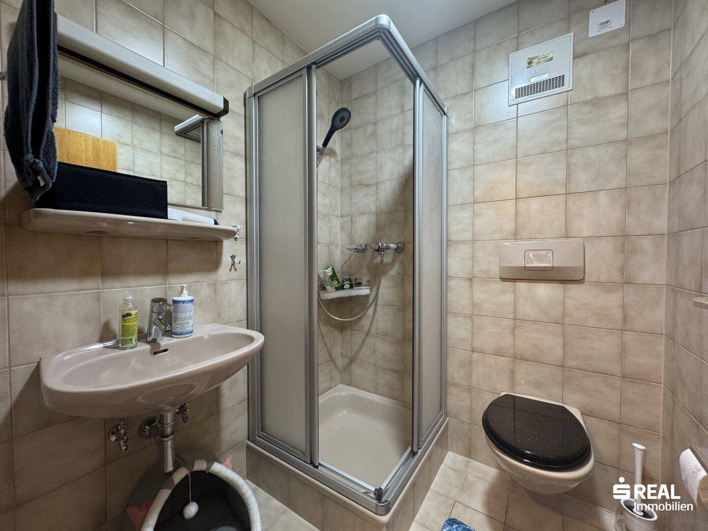Gefliestes Badezimmer mit Duschkabine, WC und Waschbecken in neutralen Farbtönen.