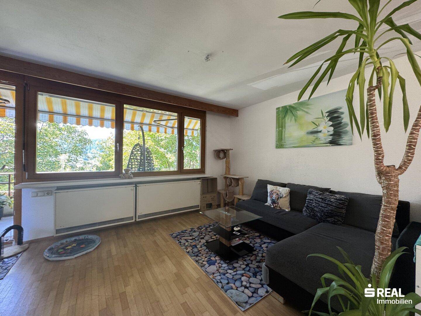 Helles Wohnzimmer mit großem Fenster zum Balkon, gemütlichem Sofa und Blick ins Grüne.