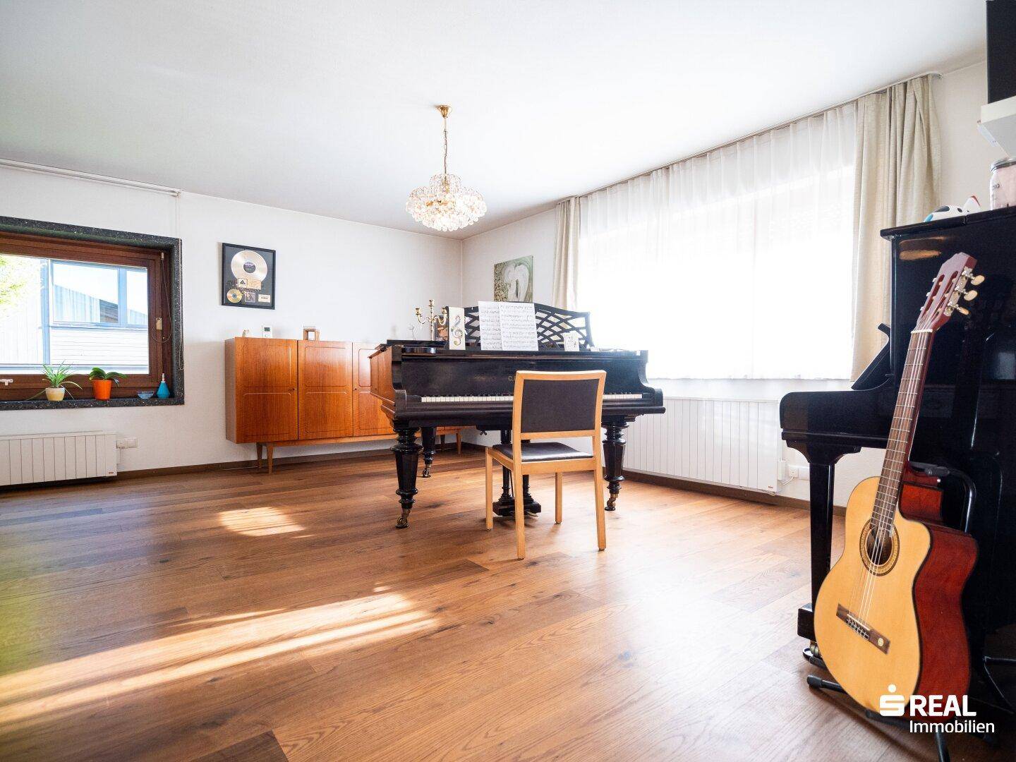 Musikzimmer mit Flügel, Klavier und Gitarre, hell durch großes Fenster.
