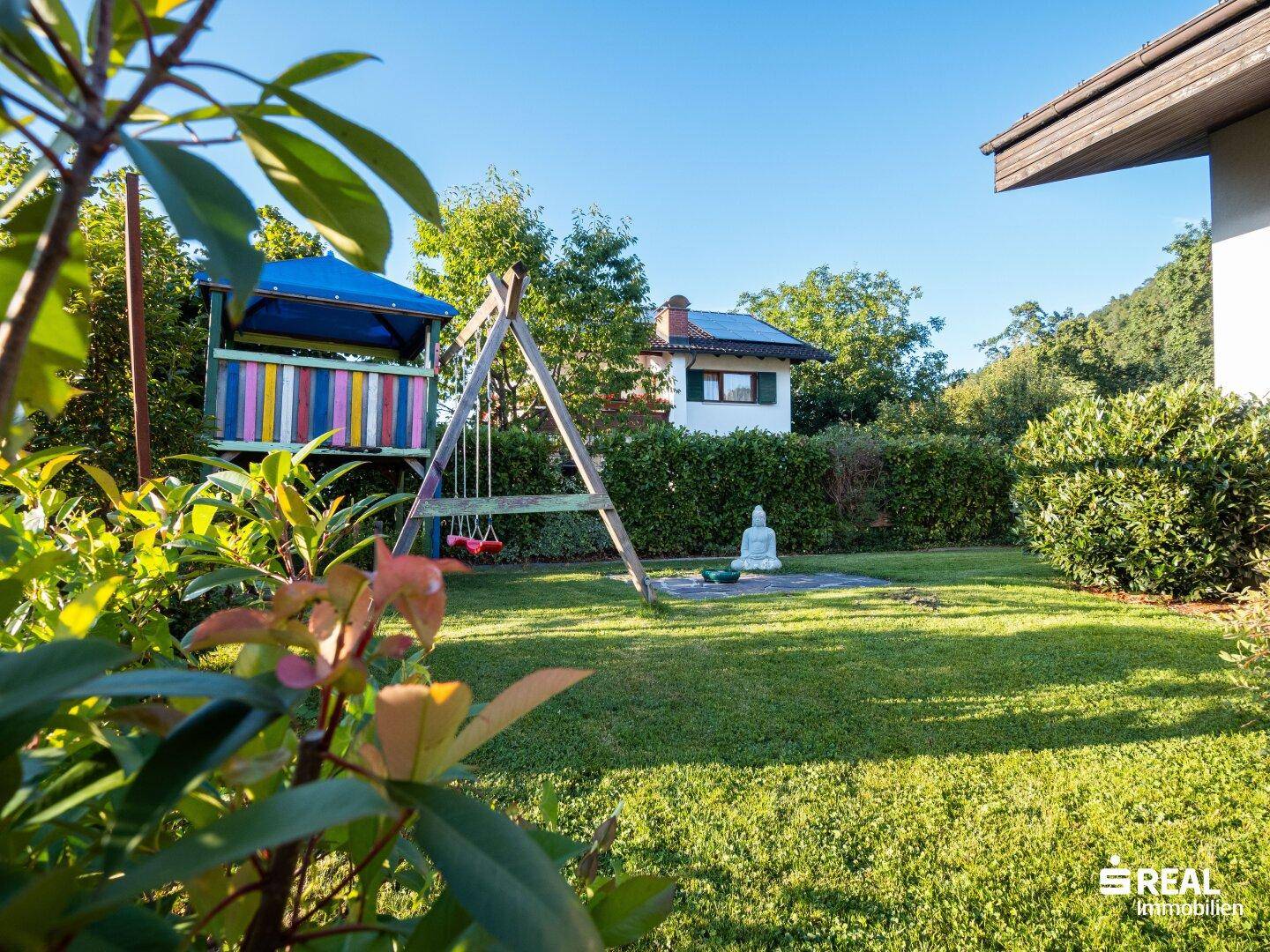 Grüner Garten mit Rasenfläche, Spielplatz und einer dekorativen Buddha-Statue.