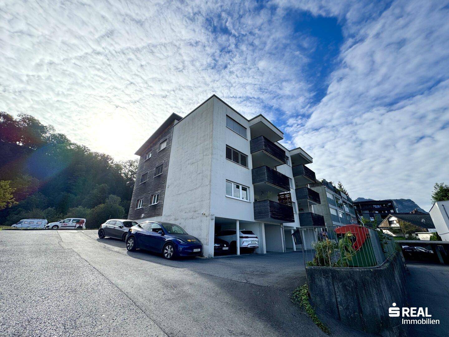 Modernes Mehrfamilienhaus mit Balkonen, Tiefgaragenstellplätzen und Bergblick unter bewölktem Himmel.