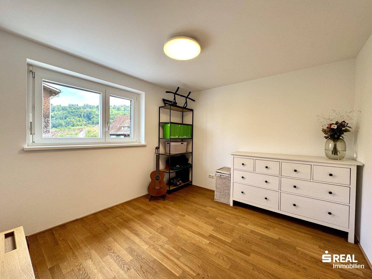 Helles Zimmer mit Holzboden, Kommode und Fenster mit Blick ins Grüne.