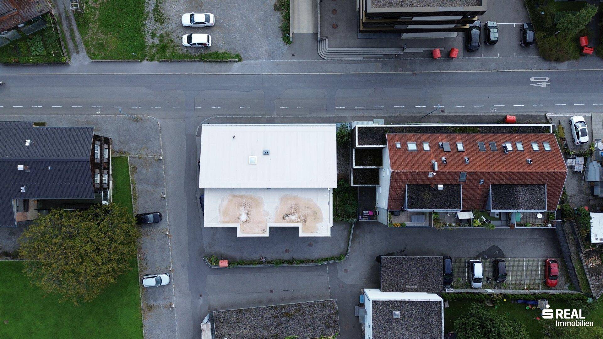 Luftaufnahme einer Wohnsiedlung mit verschiedenen Gebäuden, Dächern und geparkten Autos entlang einer Straße.