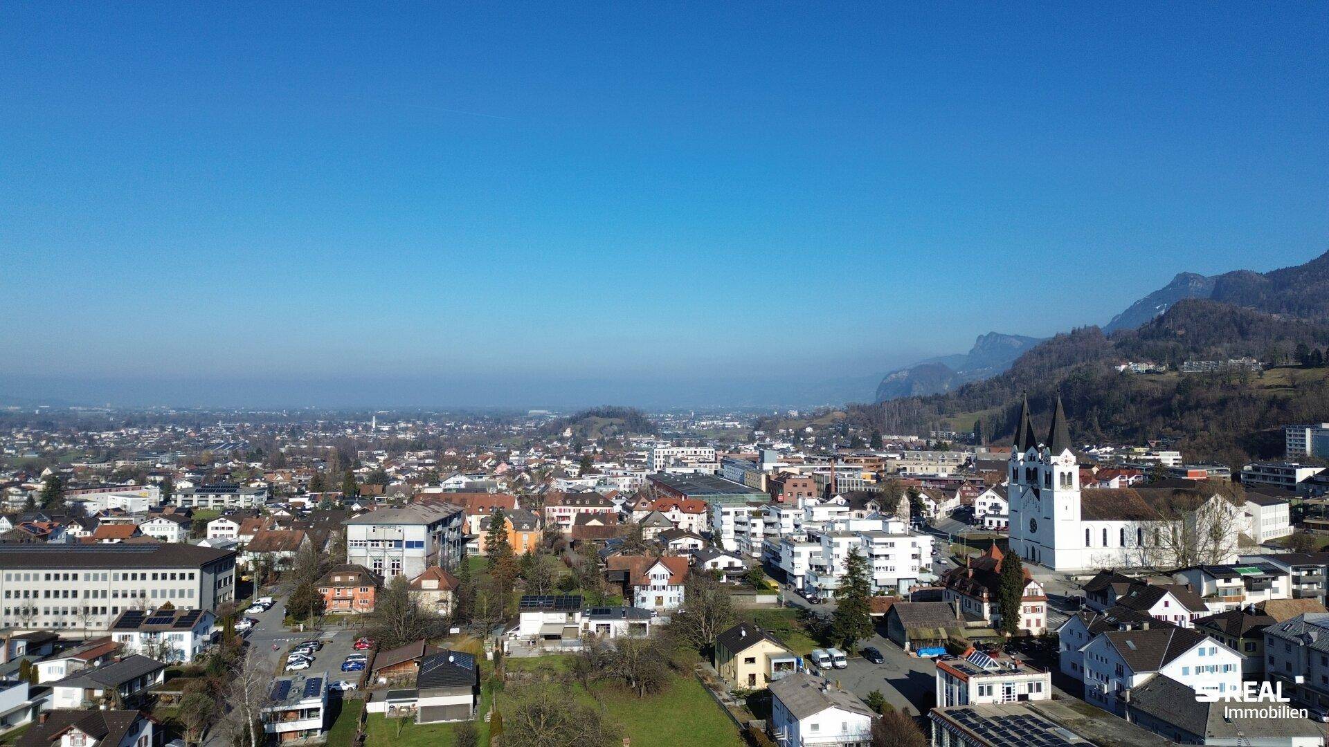 Weitläufige Luftaufnahme einer Stadt mit Wohngebäuden, Kirche und beeindruckender Bergkulisse am Horizont.