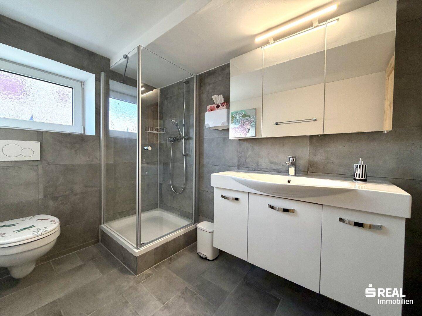 Helles Badezimmer mit Duschkabine, Waschtischunterschrank und Wand-WC, alles in modernen Grautönen.