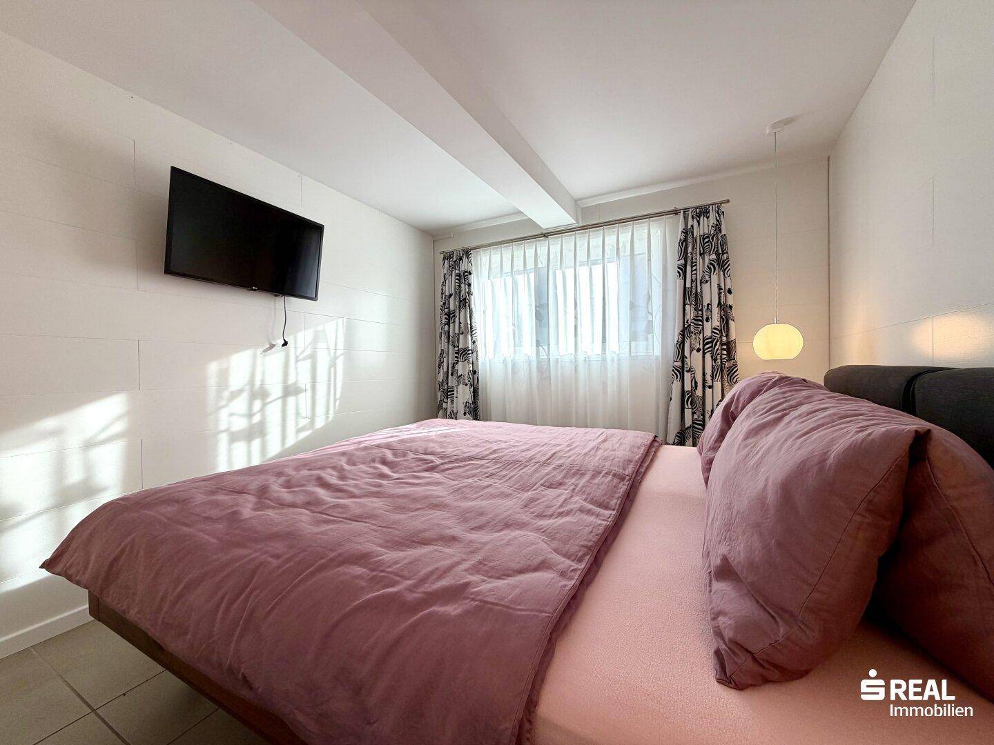 Gemütliches Schlafzimmer mit großem Bett, Wandfernseher und Fenster mit gemusterten Vorhängen.
