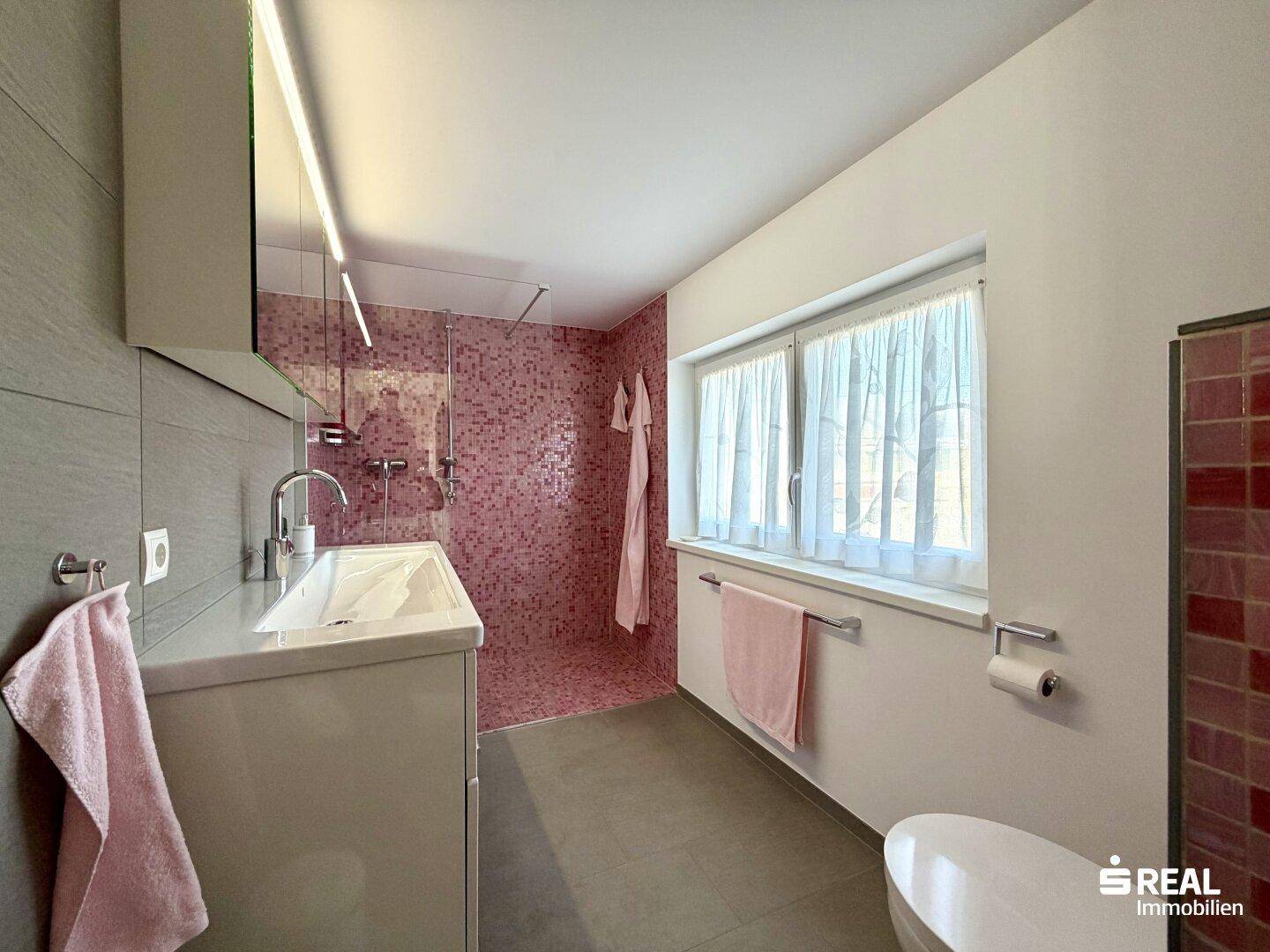 Helles Badezimmer mit Fenster, Waschtisch und begehbarer Dusche, teilweise mit Mosaikfliesen.