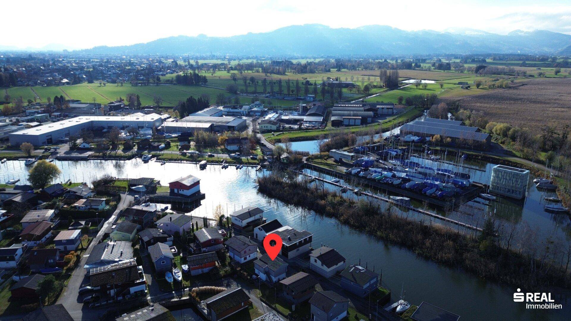 Weitläufige Luftaufnahme der Siedlung am See mit Bergen im Hintergrund und grüner Landschaft.