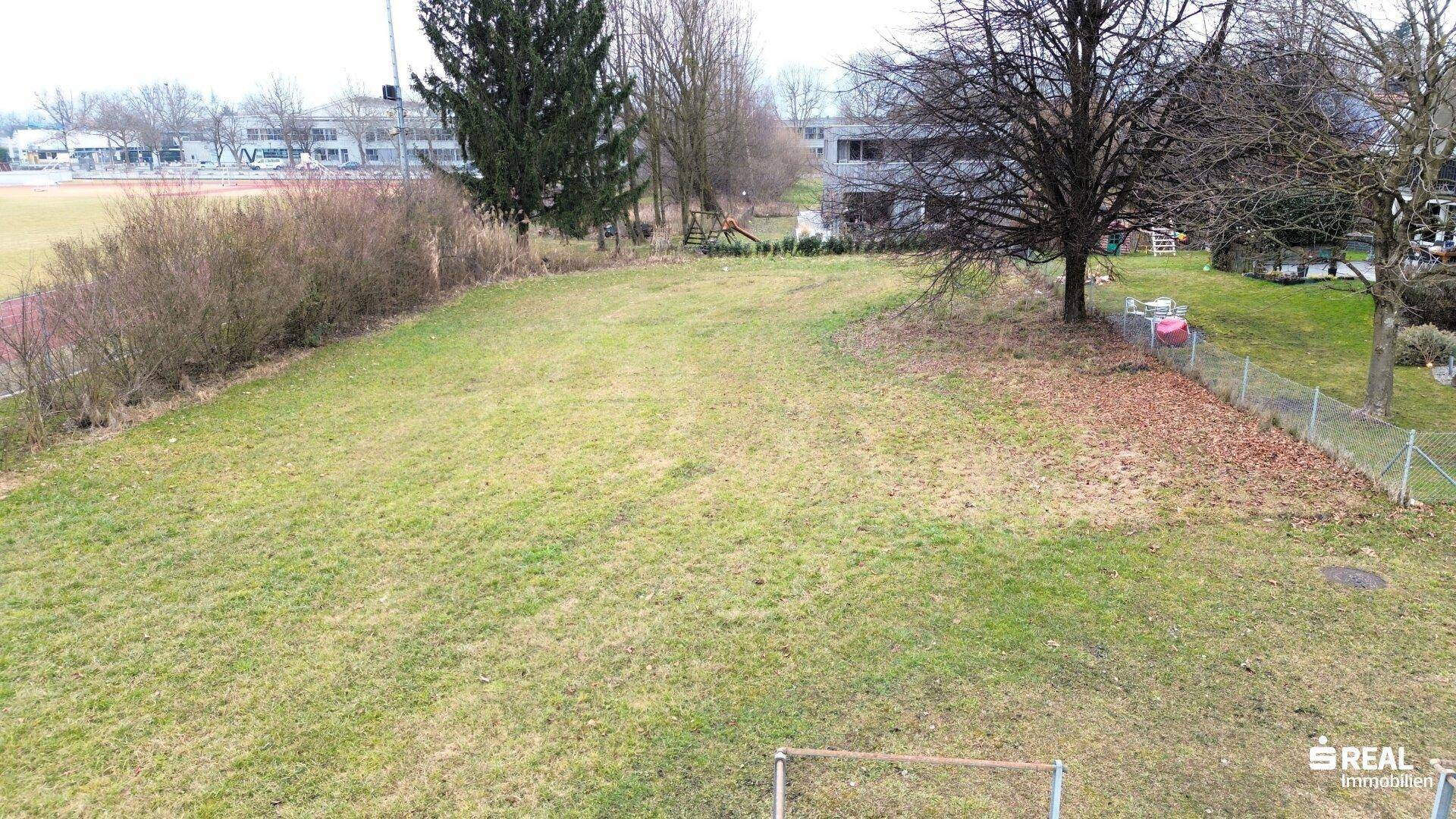 Offene Rasenfläche neben einer Sportanlage mit Laufbahn und weiteren Gebäuden im Hintergrund.