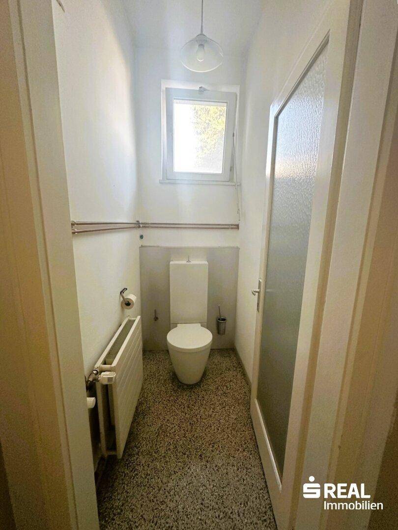 Kleine Gästetoilette mit Fenster, WC und gefliestem Boden in hellem Design und Heizkörper.