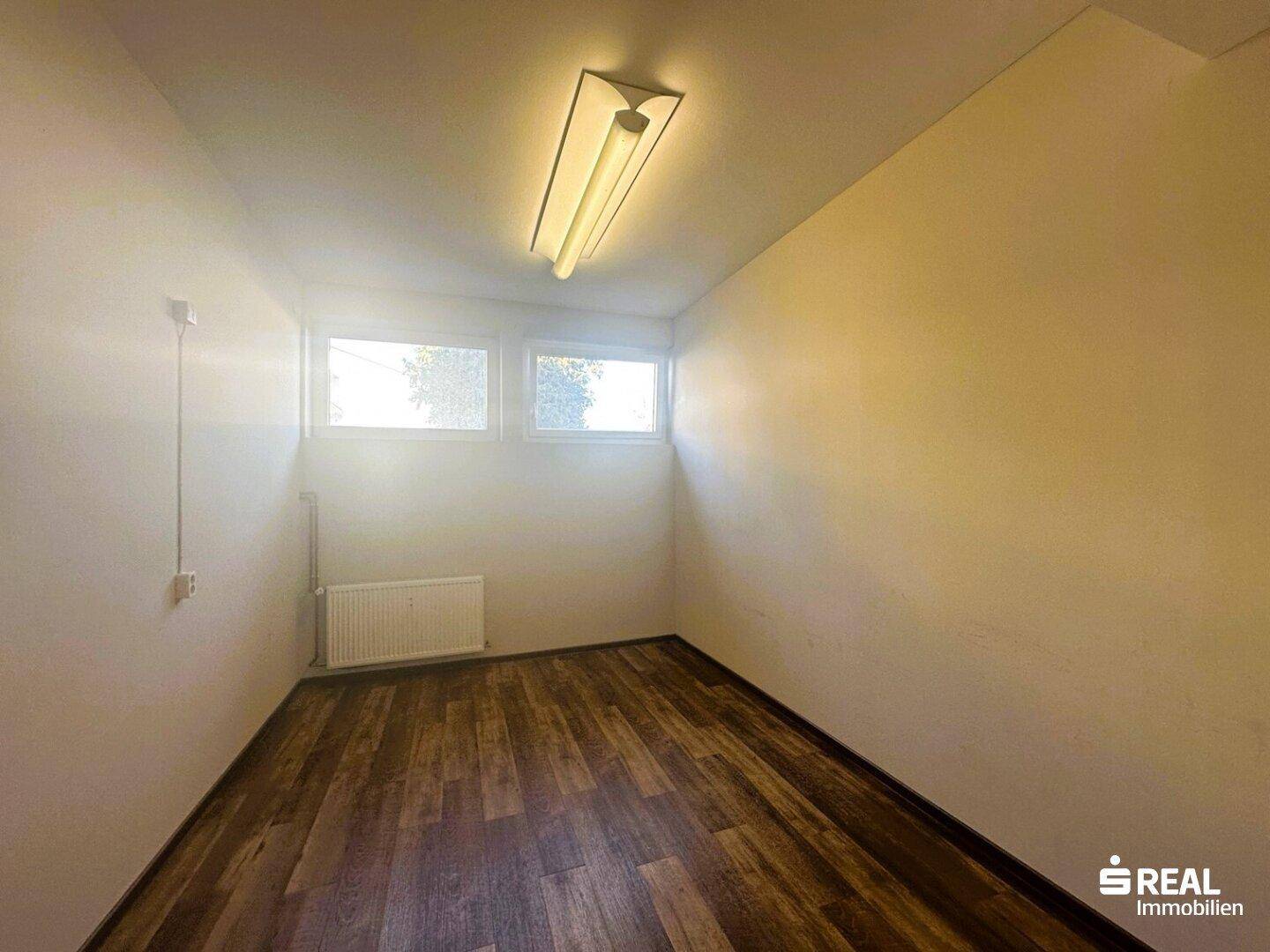 Kleiner Büroraum mit Holzboden, zwei Fenstern und einfacher Deckenbeleuchtung, ideal für konzentriertes Arbeiten.