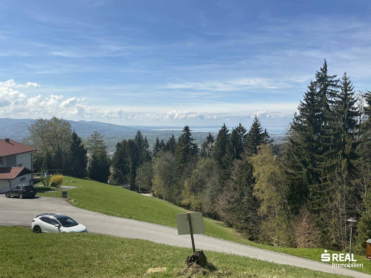 Weitläufiger Blick über eine hügelige Landschaft mit Häusern, einer Straße und einem See in der Ferne.