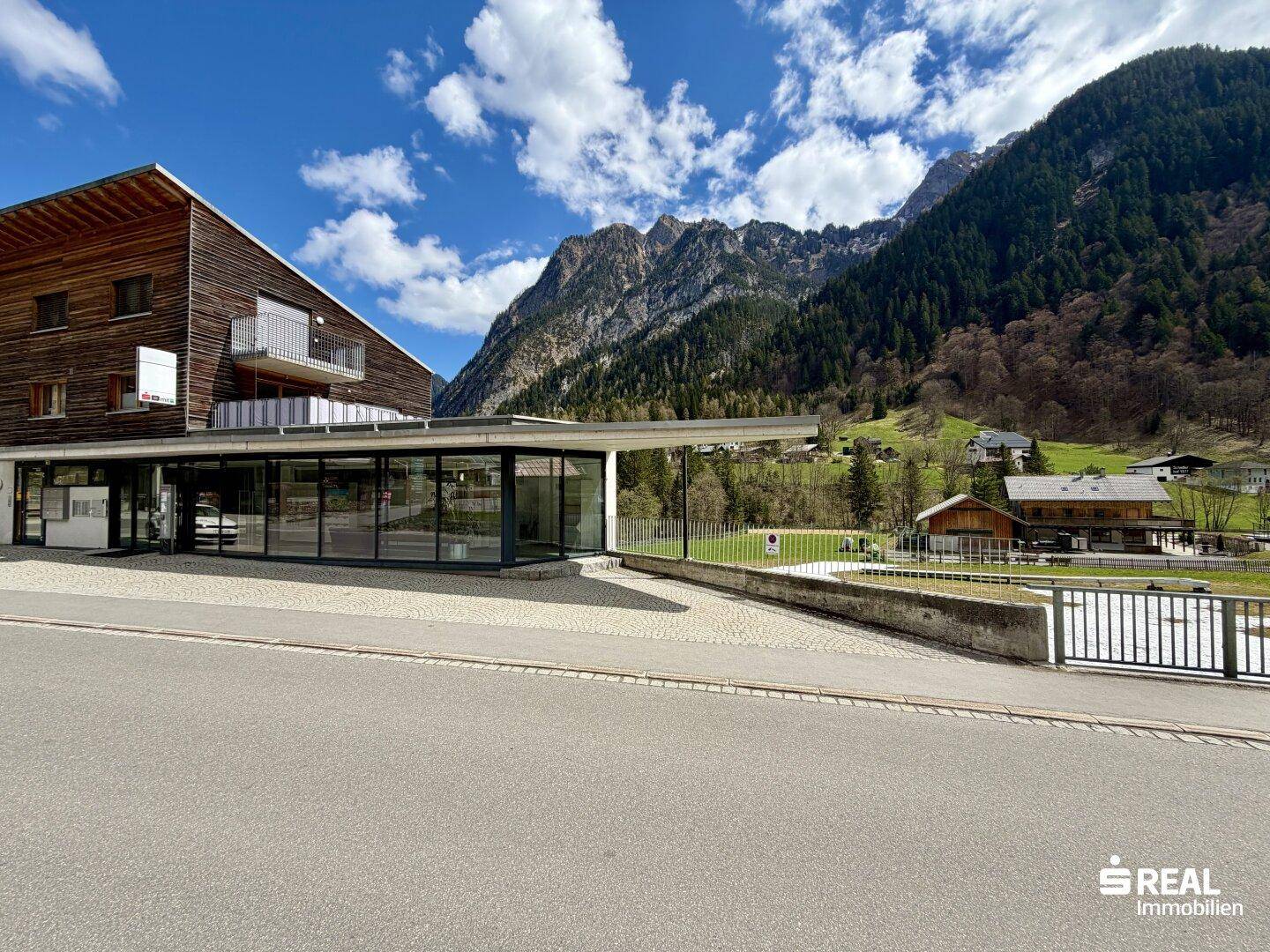 Modernes Gebäude mit Holzfassade und großen Glasflächen vor einer beeindruckenden Bergkulisse unter blauem Himmel.
