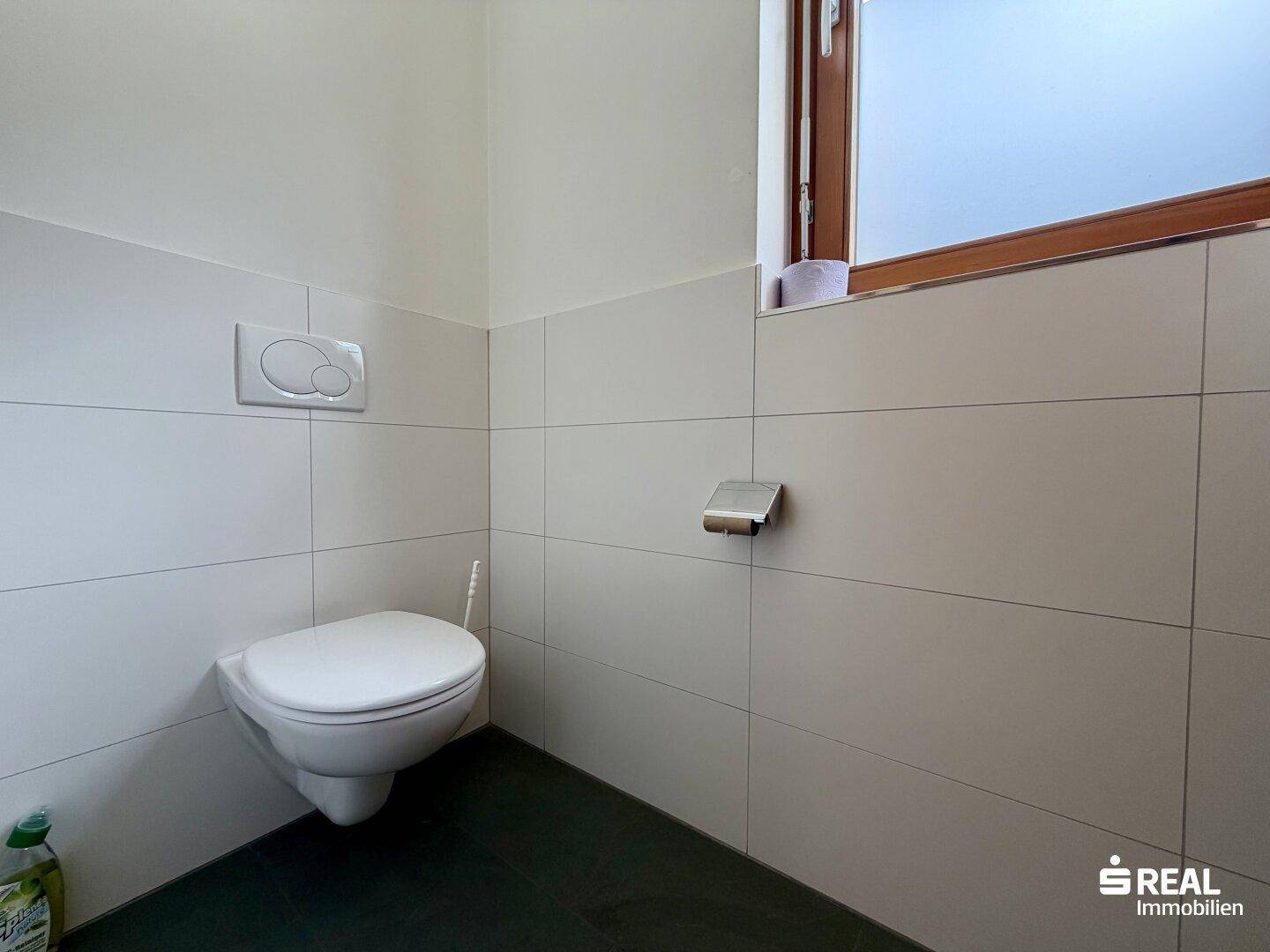 Einfache Toilette mit Wand-WC, einem Fenster und weißen Fliesen an den Wänden.